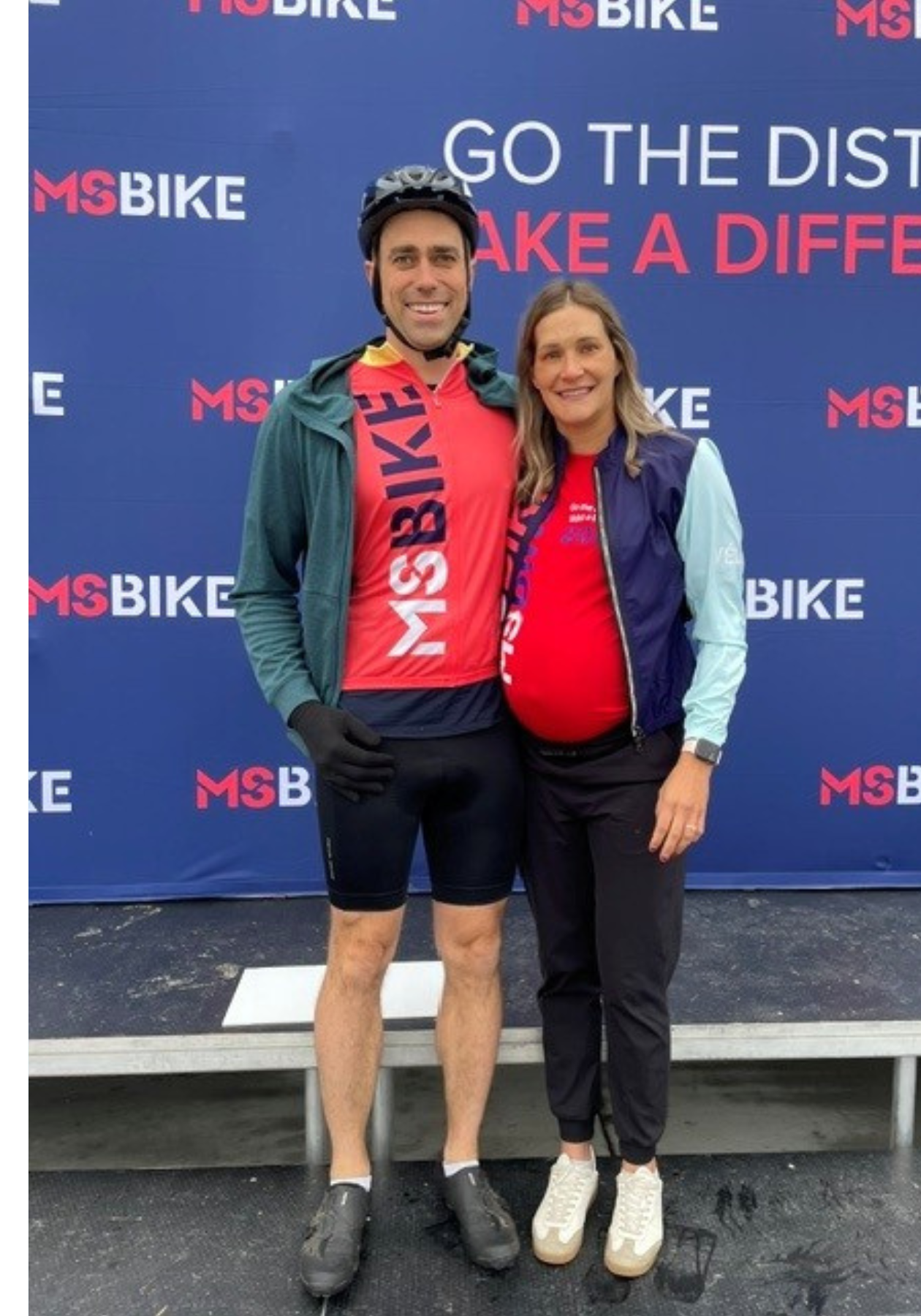 Alli and her husband wearing MS Bike jerseys posing in front of an MS Bike banner, while Alli is pregnant.